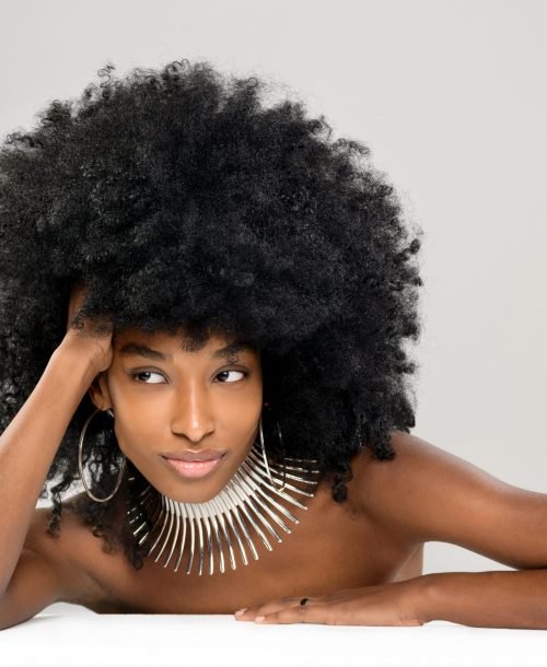 Beautiful young Black woman modelling a modern silver choker or necklace with hoop earrings leaning over a table with her bare shoulders looking aside with a quiet smile